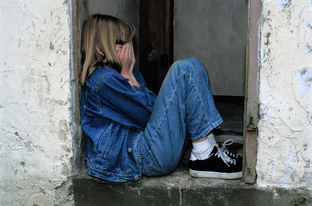 ein Mädchen sitzt auf einer Mauer und hält sich mit den Händen das Gesicht verdeckt und trägt Jeans mit Turnschuhen